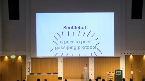 A picture of Zenna Fiscella on stage giving a talk; she is facing the audience, and behind and above her is a projected screen approximately 10 meters wide showing a slide that says: Scuttlebutt – a peer to peer gossiping protocol