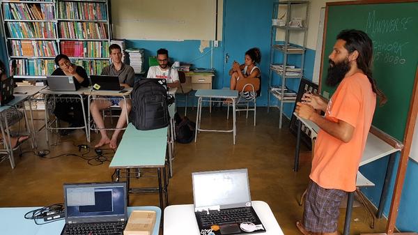 In a small Brazilian school classroom, there are a dozen people at desks with their computers open while listening to Luandro speaking in front about SSB, Manyverse, and Patchwork