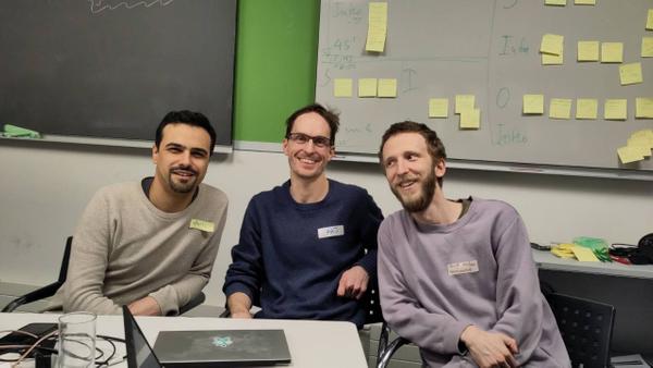 A photograph of staltz, arj, and powersource sitting on chairs around a desk, smiling