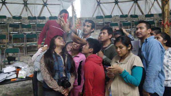 In a small auditorium that seems to be a tent, there are a dozen young university students holding their phones up in selfie position or using it in normal stance; some are smiling, some are focused on exploring their phones