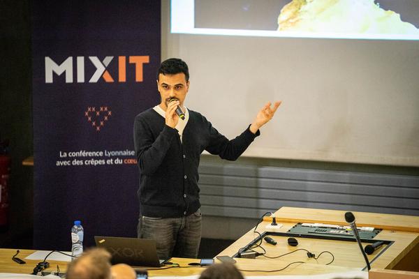 Photograph of Andre Staltz holding a microphone while giving a talk; behind him you can see a vertical poster of the Mixit conference, and a part of the projected screen where slides are shown