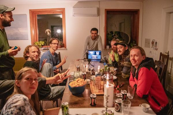 Photograph of 8 people sitting and standing around a rectangular table, indoors, in the evening. On the table, laptops, snacks, drinks. People are looking at the camera and smiling